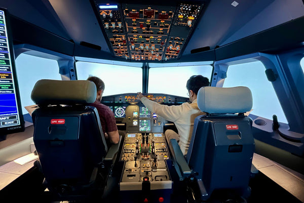 Two people operating a realistic commercial flight simulator cockpit with illuminated panels.