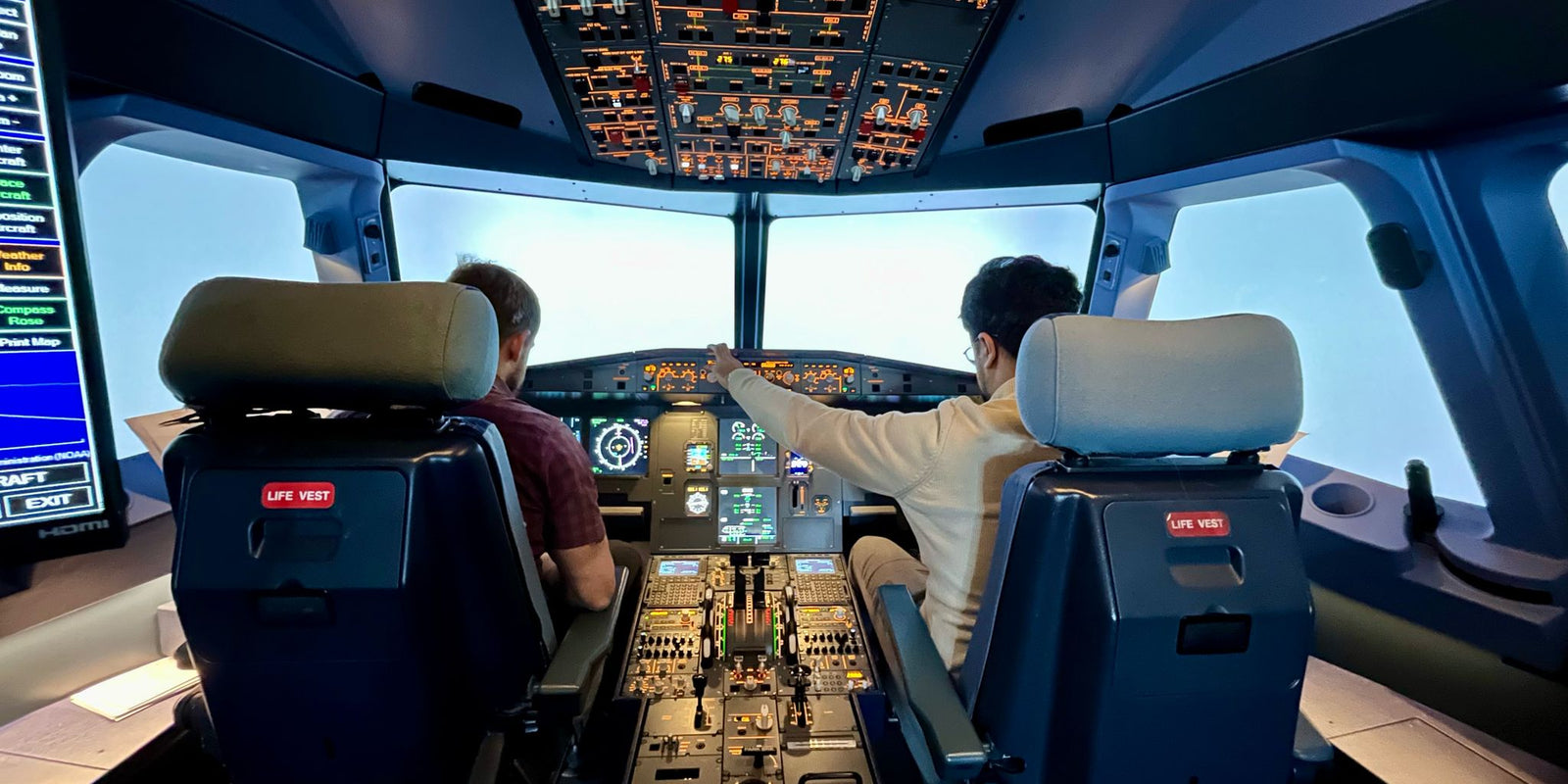 Two people operating a realistic commercial flight simulator cockpit with illuminated panels.